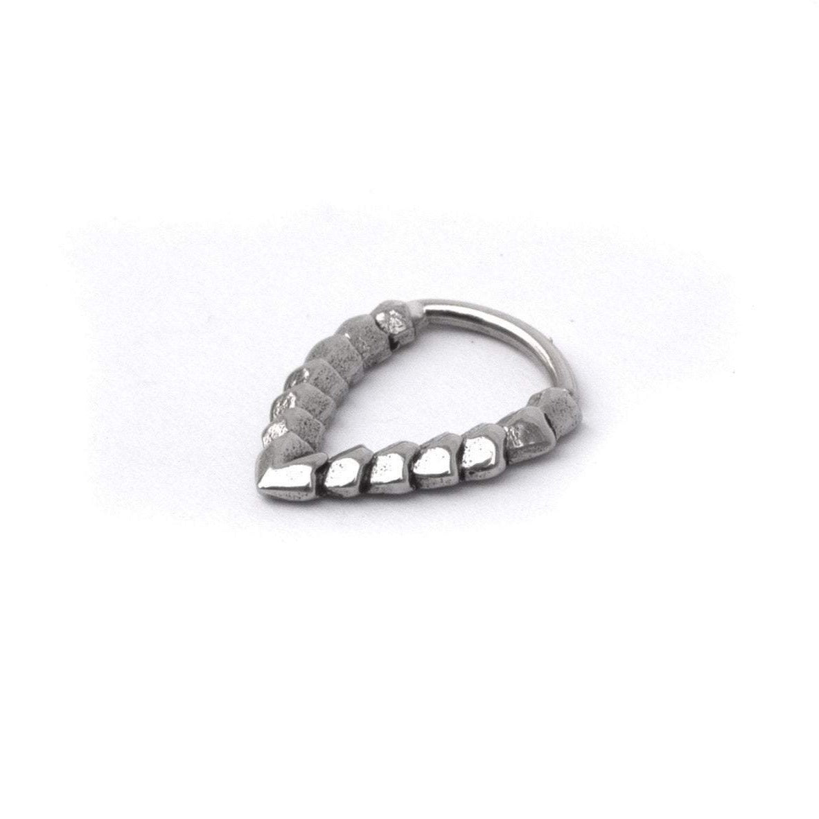 The Kaya Movement KAMO Biomech Teardrop Clicker Septum Ring in Silver | 16 gauge is a uniquely designed, handcrafted piece of body jewelry. It features a sequence of small geometric shapes that create a gradient-like pattern, starting with smooth, rounded forms on one end and transitioning to textured, angular shapes on the other, forming an elegant teardrop shape.